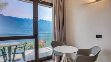 Un balcón con una mesa y sillas, vista a un lago y montañas, y una cortina en la ventana.