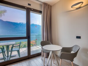A balcony with a table and chairs, a view of a lake and mountains, and a curtain on the window.