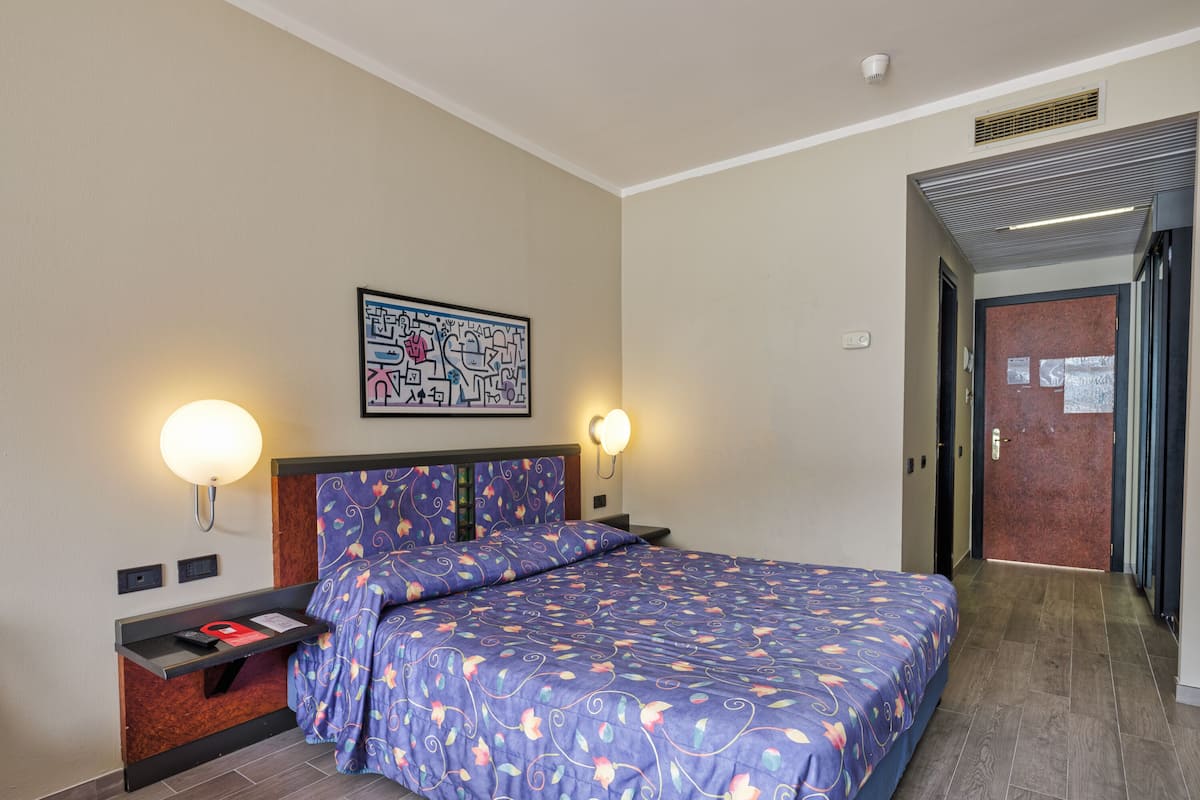 A hotel room with a bed, two bedside lamps, a wall-mounted artwork, and a door leading to another room.