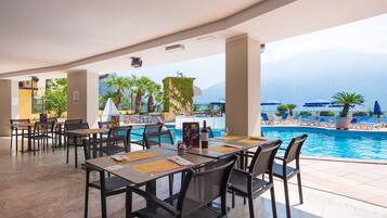 Un restaurante junto a la piscina con mesas al aire libre, piscina y vistas a montañas.