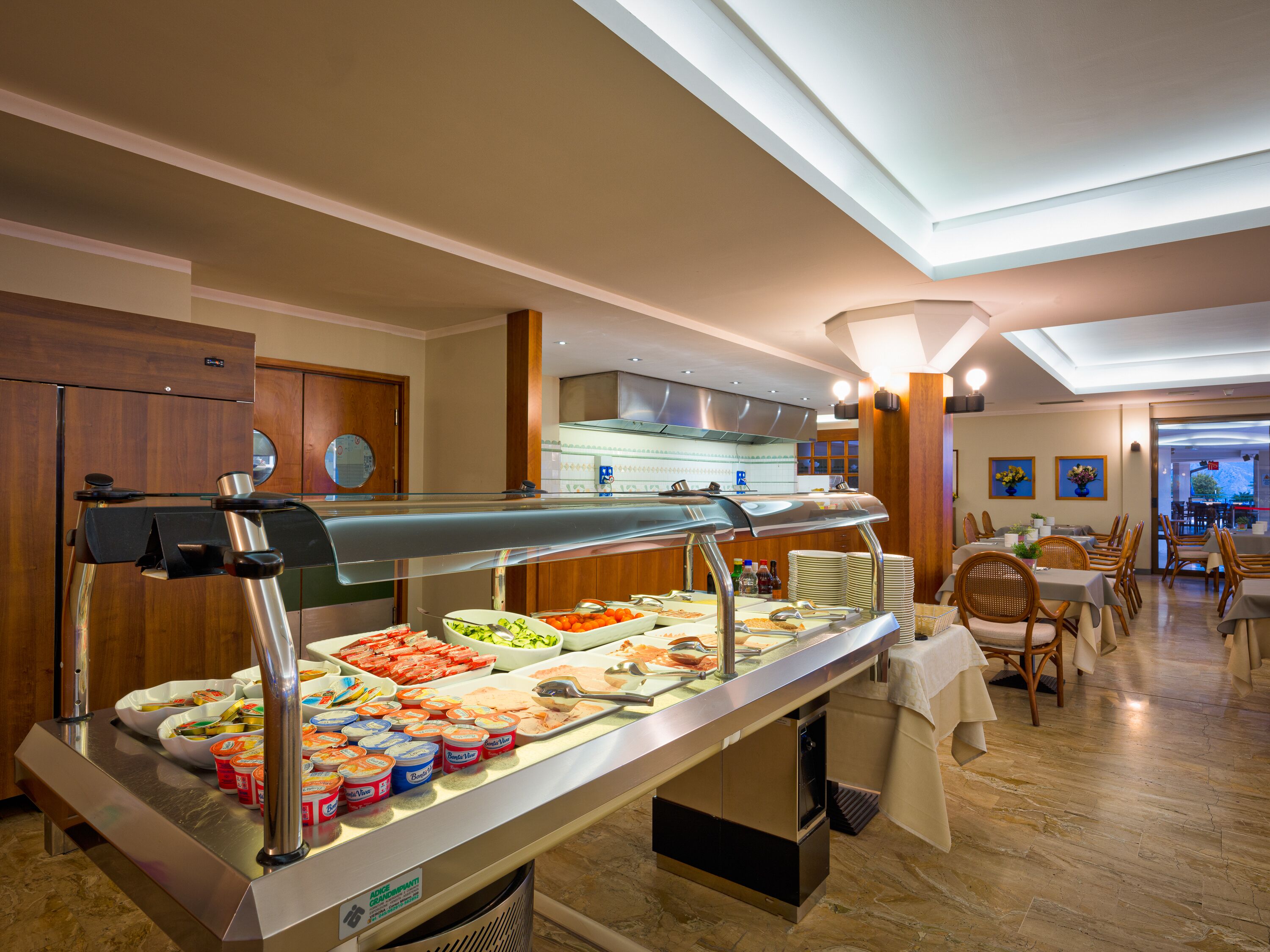 Zona de bufé con diversas opciones de comida, una cocina al fondo y un comedor con mesas y sillas.