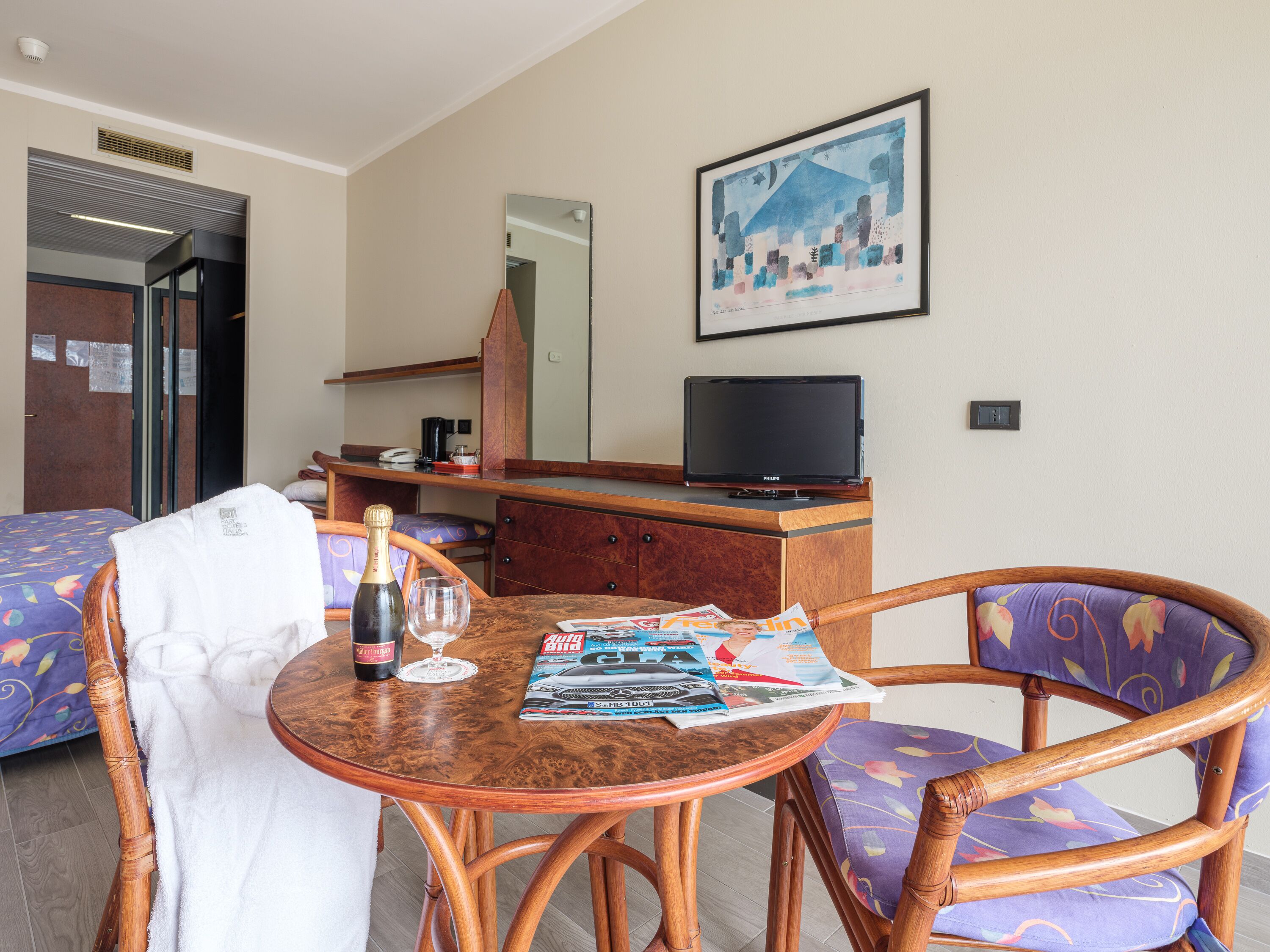 Ein Hotelzimmer mit einem Holztisch, zwei Stühlen, einem Fernseher, einer Champagnerflasche und einer Zeitschrift.