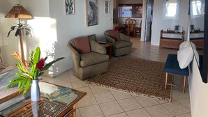 A living room with a sofa, armchairs, a coffee table, and a rug. There are framed pictures on the wall, a lamp, and a plant on the floor. - Maui Beach House (Lahaina)