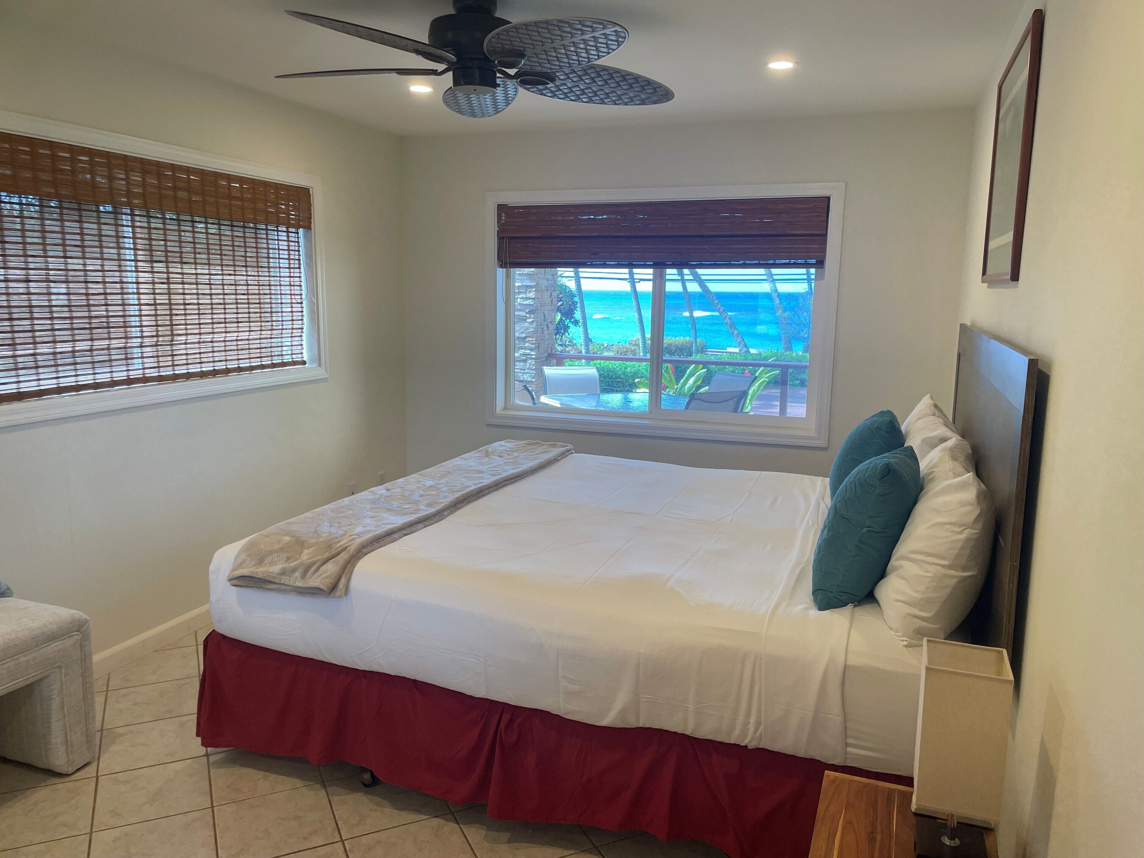 A bedroom with a bed, a ceiling fan, a window with a view of the ocean, and a small table with a lamp.