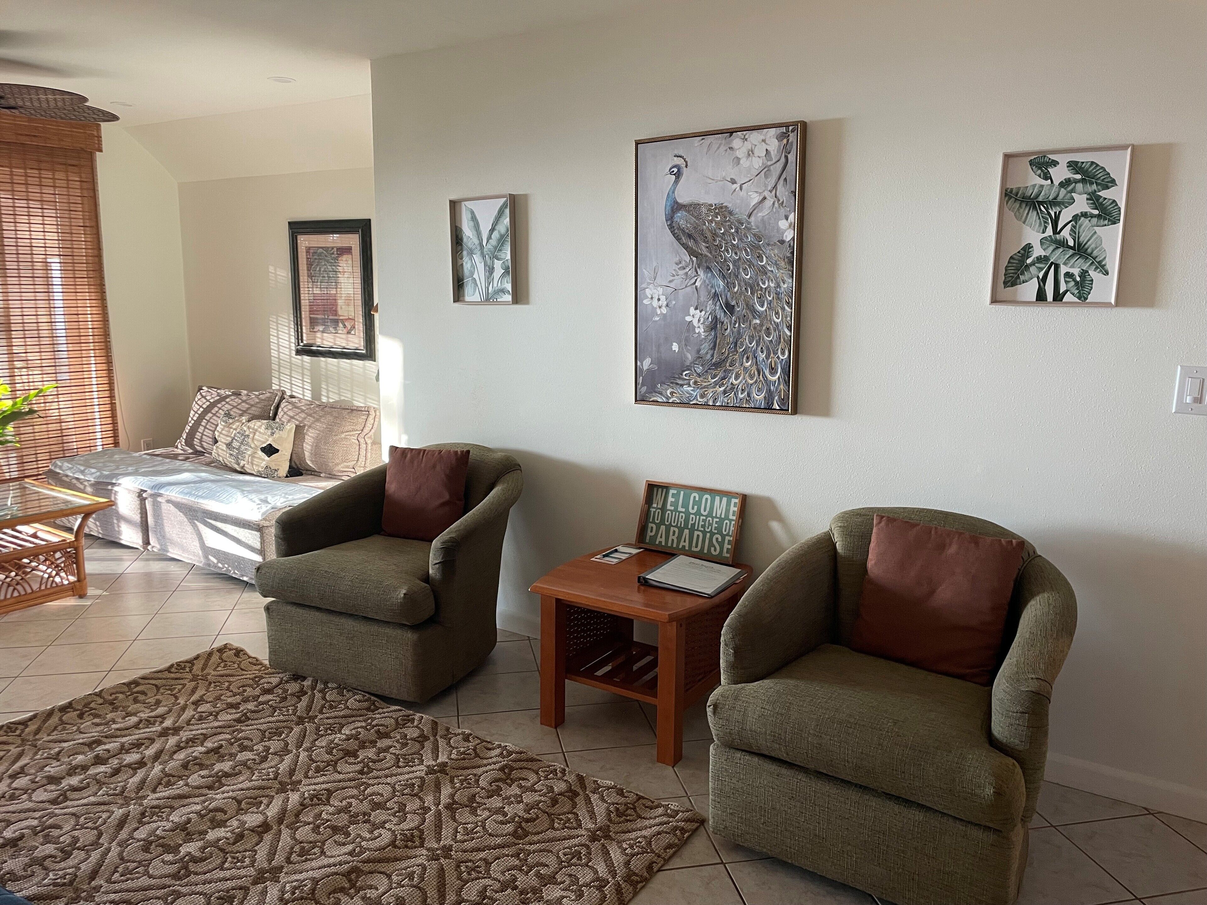 Un salon comprenant deux fauteuils, un canapé, une table basse et un tapis à motifs. Les murs sont ornés d’œuvres d’art représentant un paon et du feuillage.