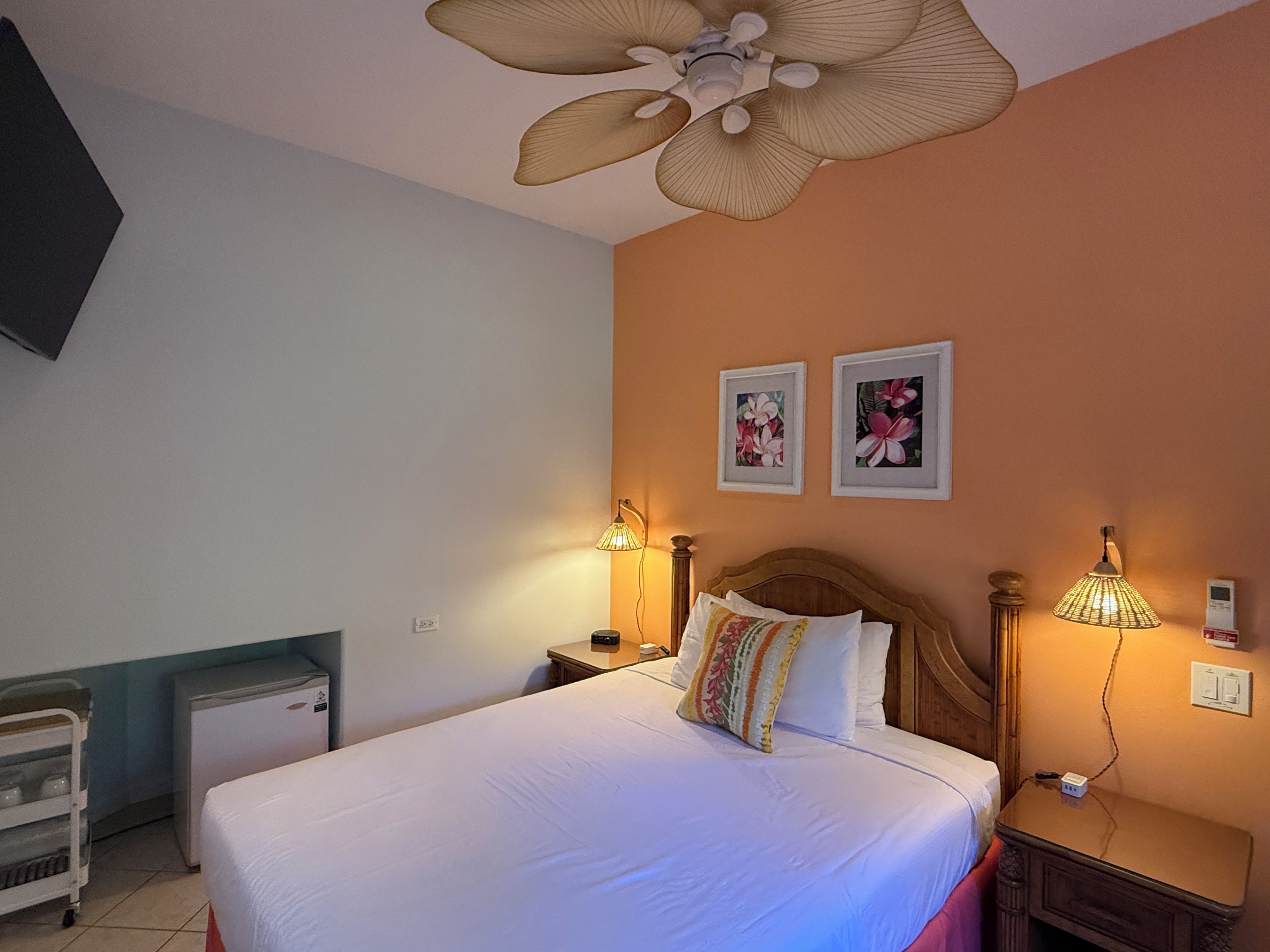 Quarto de hotel com cama, abajures, ventoinha no teto, pequeno frigorífico e duas quadros na parede.