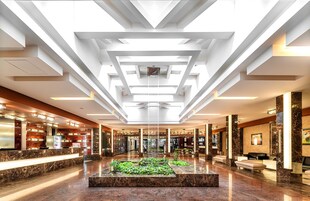 Um hall amplo com design moderno no teto, um espelho d'água central e uma área de recepção com balcão de mármore.