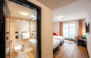 Quarto de hotel moderno com porta de vidro para um banheiro, cama com cabeceira vermelha, televisão e escrivaninha com cadeira.
