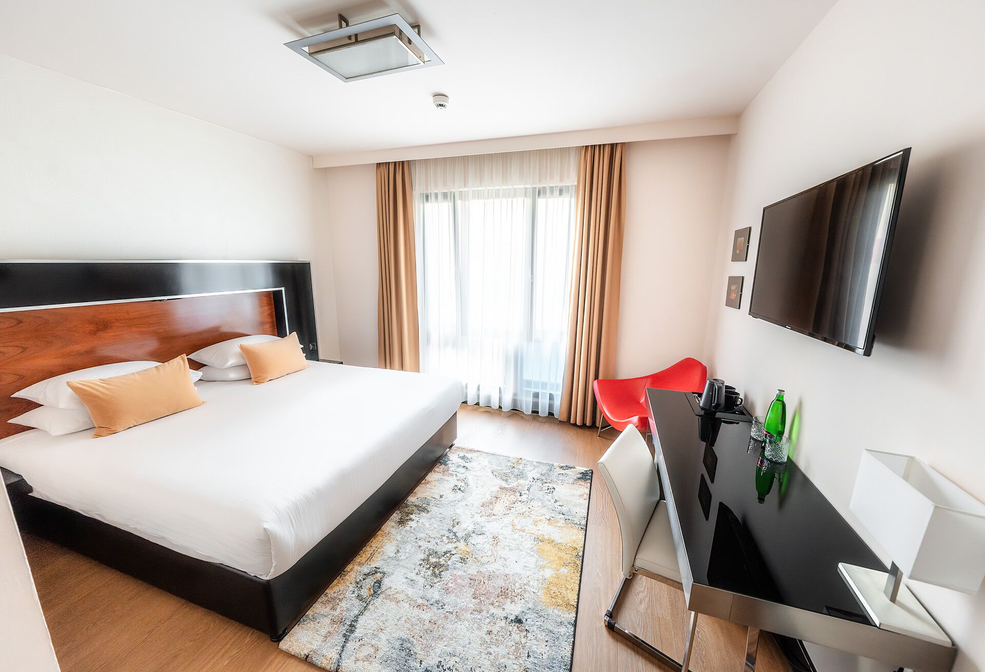 A modern hotel room with a large bed, a desk, a flat-screen TV, and a window with curtains.
