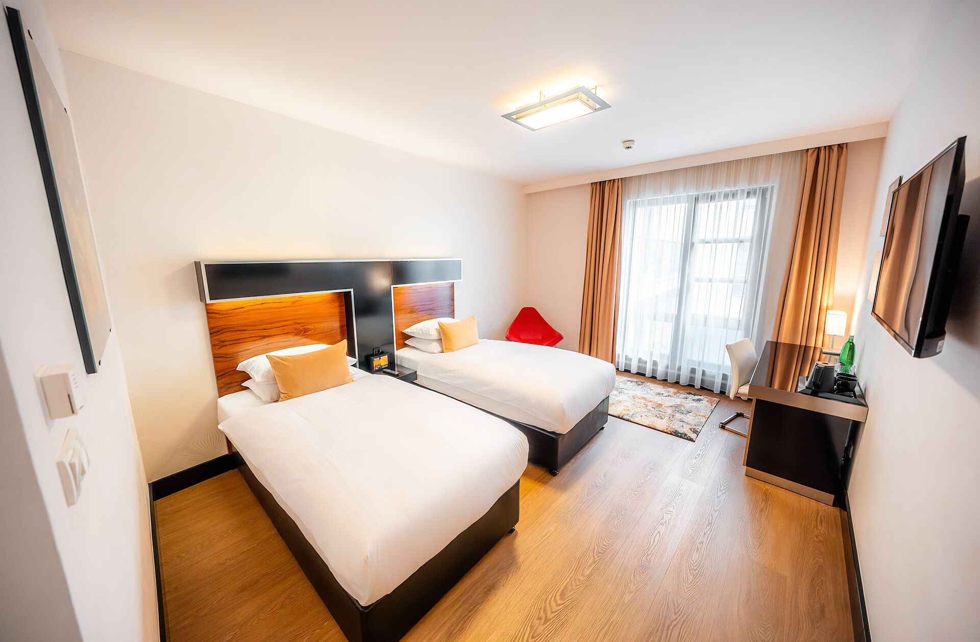 Ein Hotelzimmer mit zwei Betten, einem roten Sessel, einem Schreibtisch mit Lampe und einem großen Fenster mit Vorhängen.