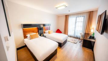 A hotel room with two beds, a red chair, a desk with a lamp, and a large window with curtains.
