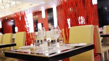 Una mesa de comedor dispuesta con copas de vino, cubiertos y una servilleta, con sillas y un elemento decorativo rojo al fondo.