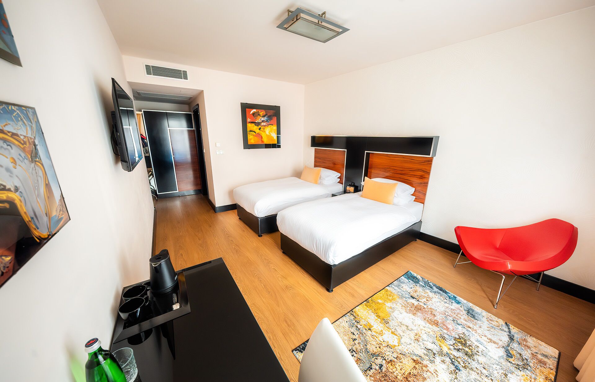 Una habitación de hotel con dos camas, una silla roja, una cafetera y obras de arte en las paredes.
