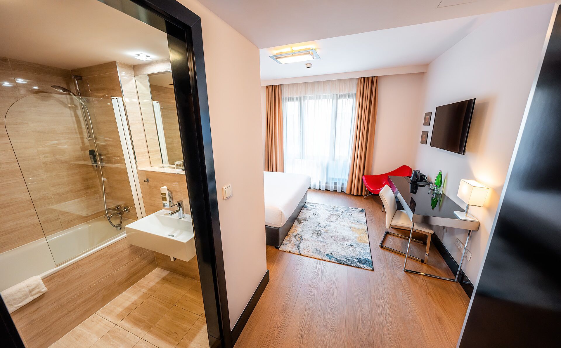 A modern hotel room with a shower, sink, and a dining area with a TV and table.