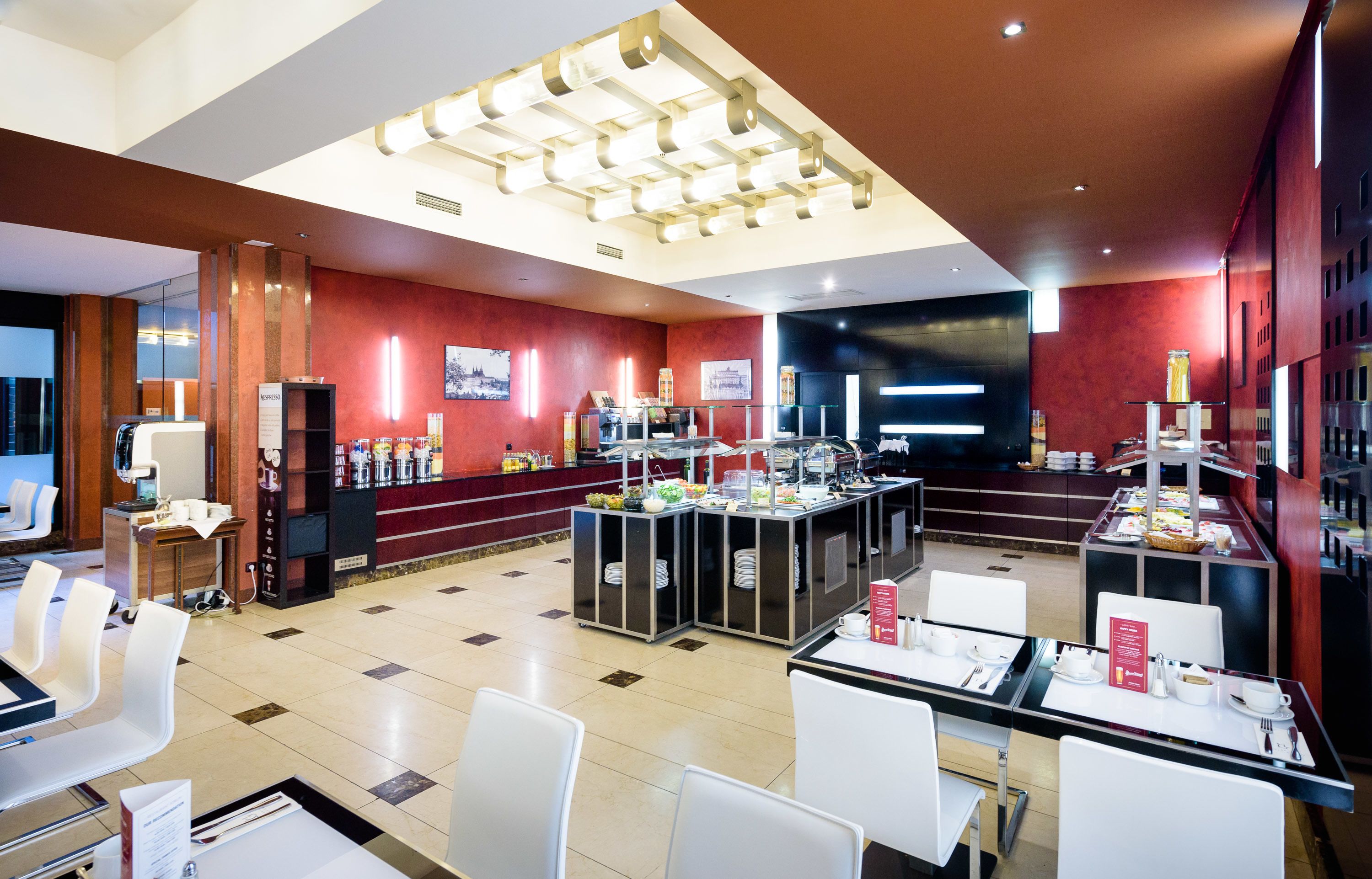 Un comedor moderno de hotel con varias estaciones de buffet, sillas blancas y un interior limpio y bien iluminado.