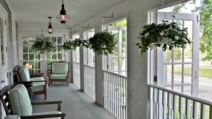 A covered porch with white railings, green cushioned chairs, hanging flower baskets, and pendant lights. - Mill House Inn (East Hampton)