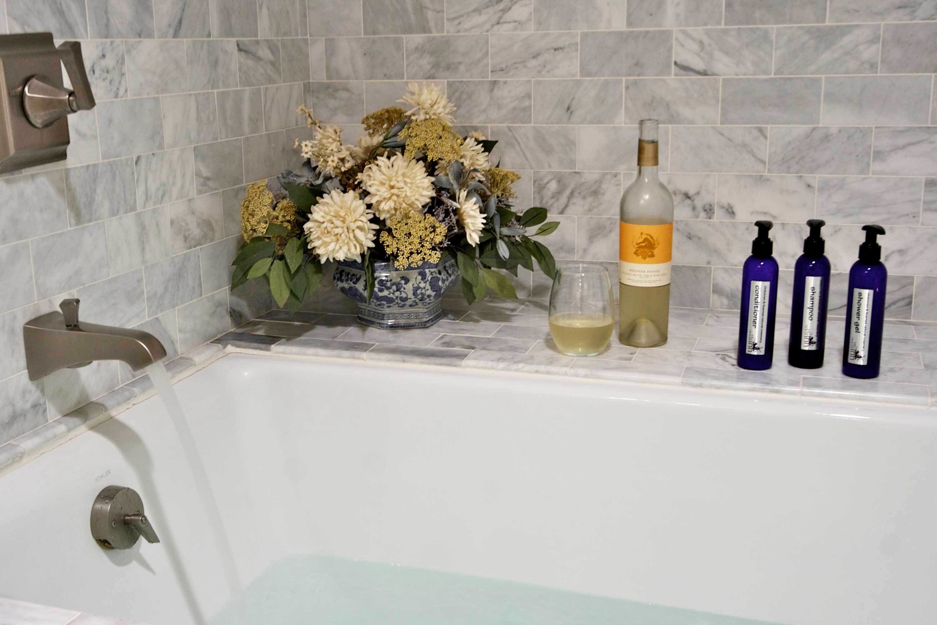 A bathroom with a marble tiled wall, a bathtub, a vase with flowers, a bottle of lotion, and a glass of water.