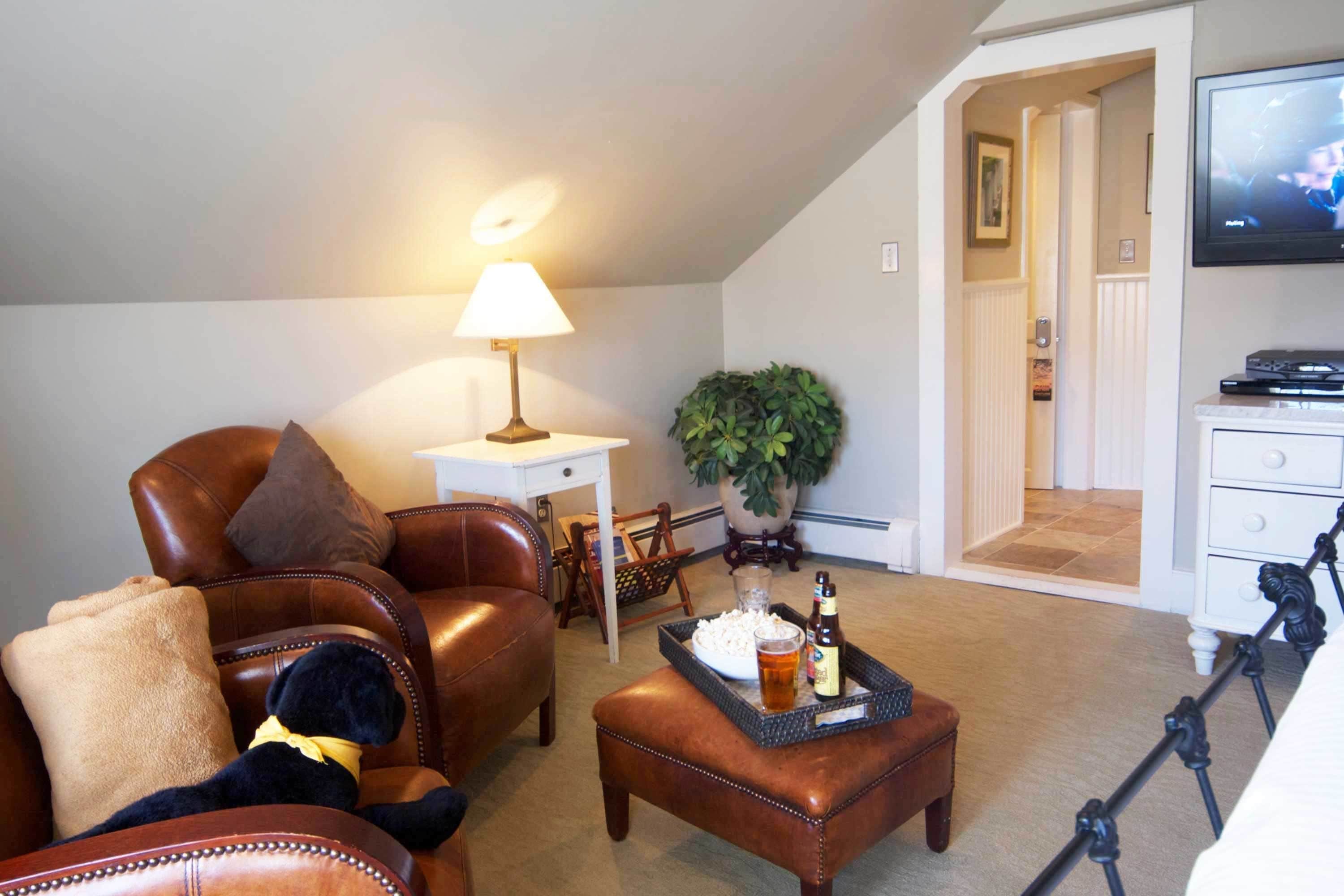 A cozy living room with a leather sofa, a coffee table with a tray of snacks and drinks, a lamp, and a television on the wall.