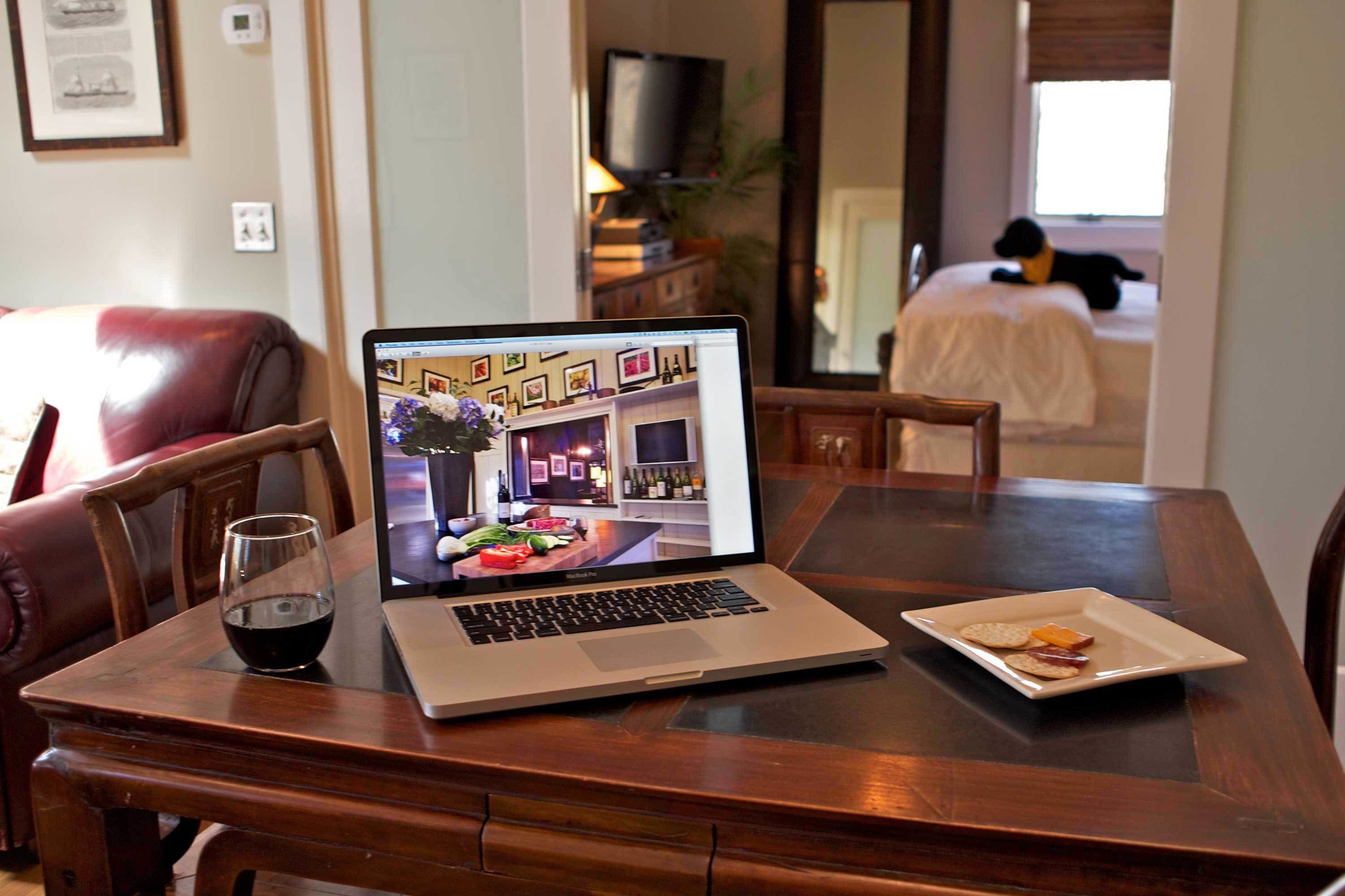 A laptop on a wooden table with a glass of red wine and a plate of food.