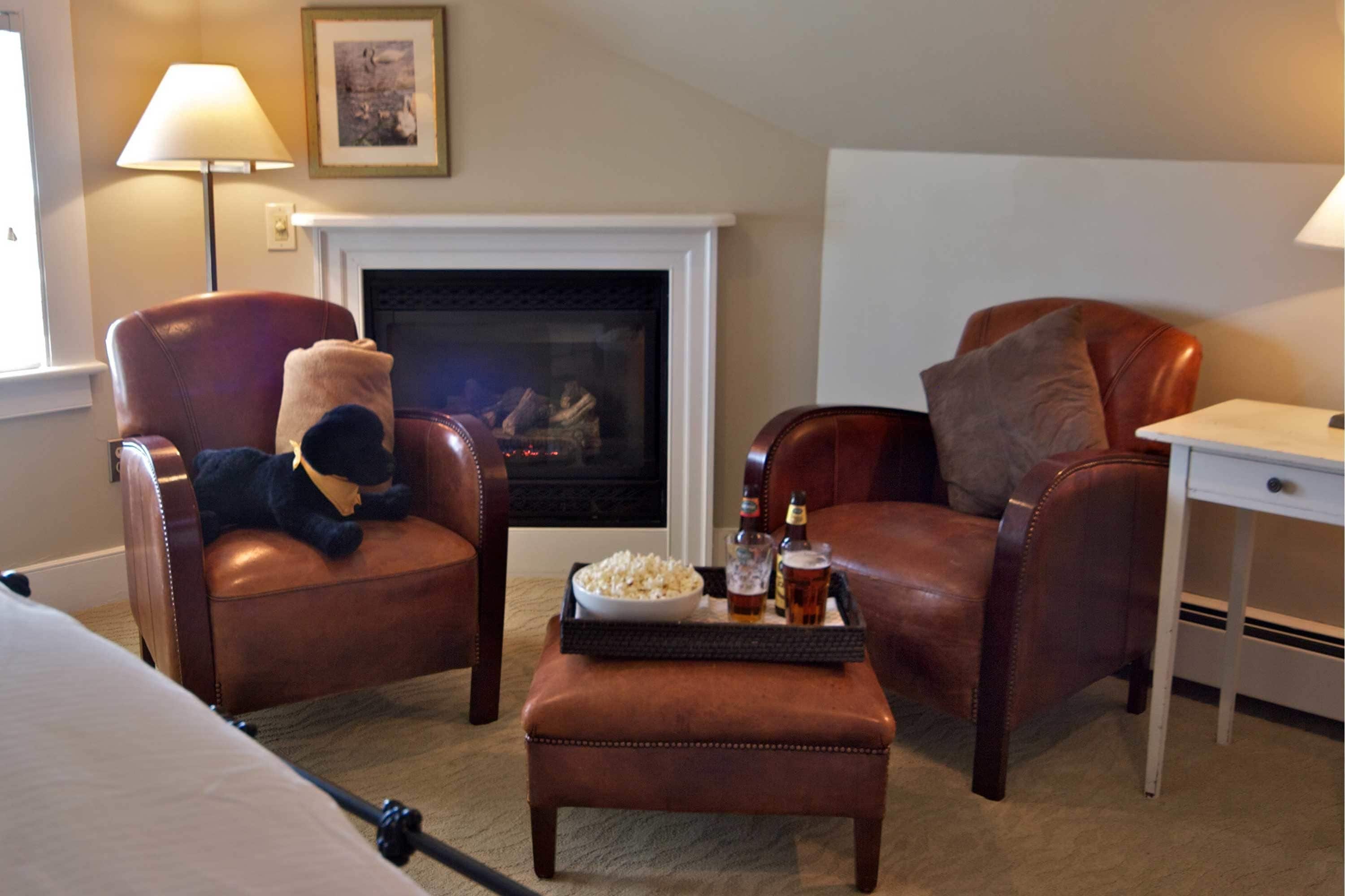 A hotel room with two leather armchairs, a small ottoman, a tray with snacks and drinks, a fireplace, and a desk with a lamp.