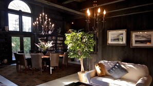 A cozy living room with a white sofa, a dining table with chairs, a chandelier, a potted plant, and framed pictures on the wall. - Mill House Inn (East Hampton)