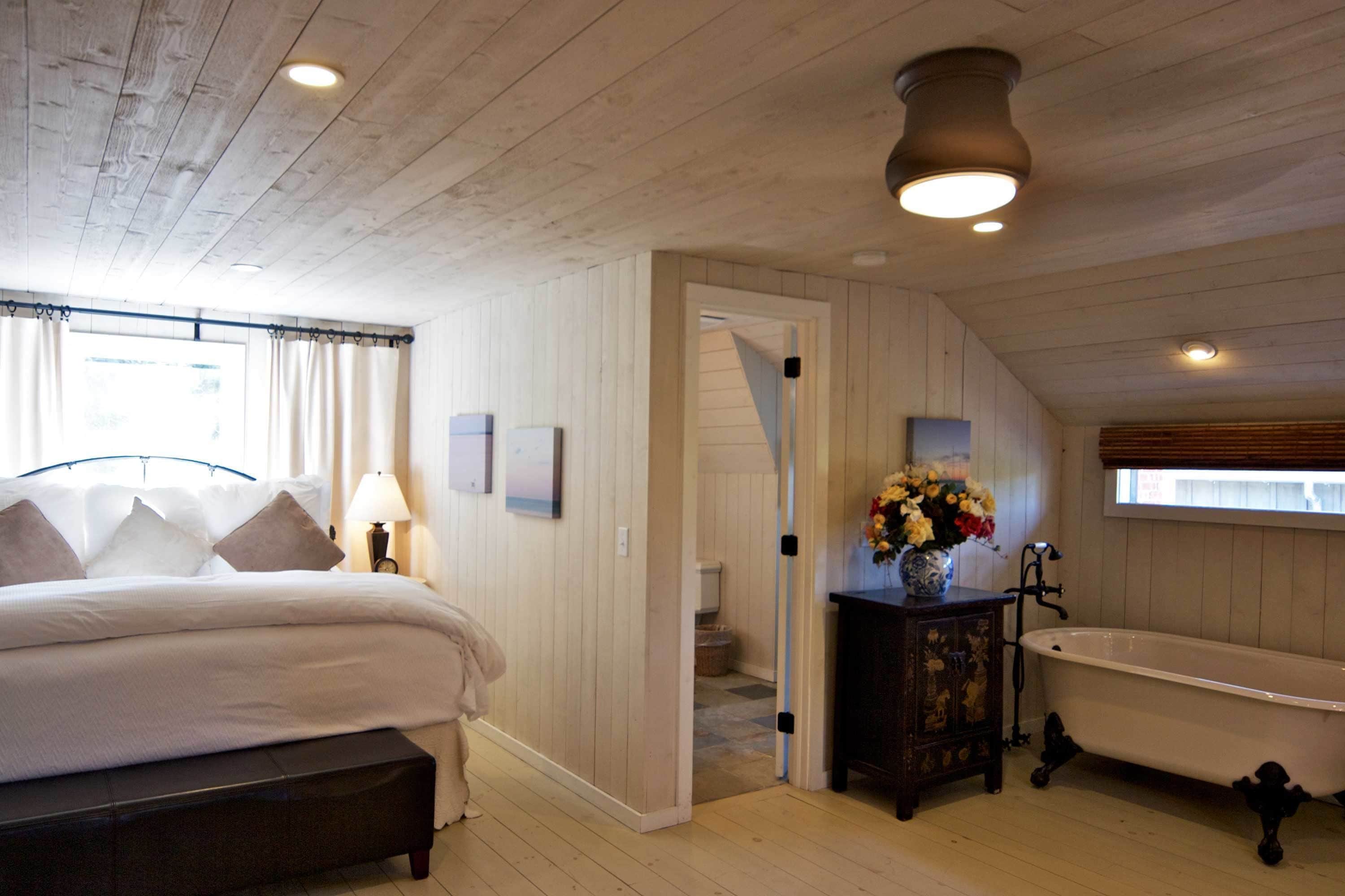Un dormitorio con cama, mesita de noche con lámpara, ventana con cortinas, techo de madera y un baño con bañera.