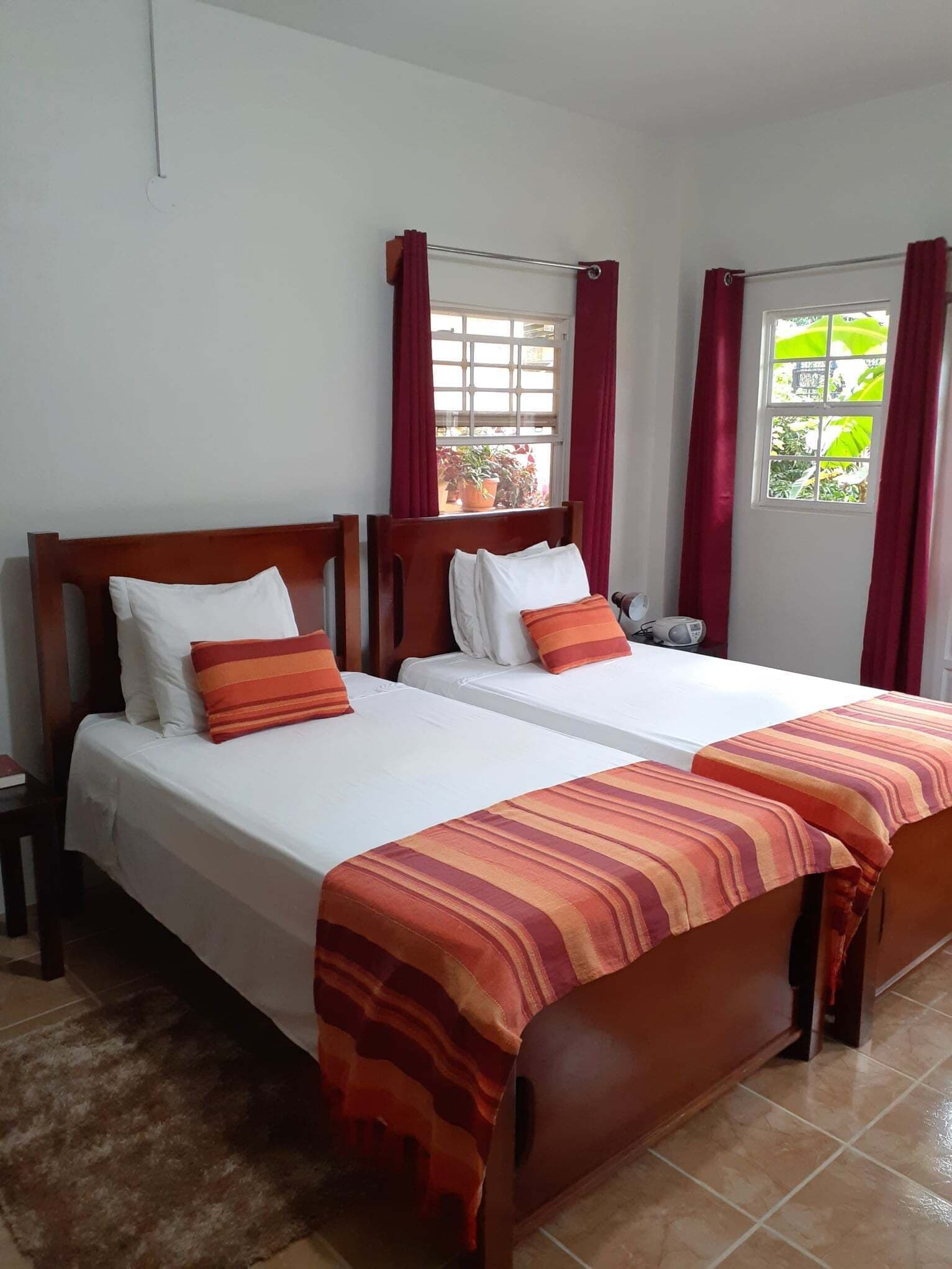 Habitación doble con dos camas, cada una con ropa de cama blanca y colchas a rayas anaranjadas. Hay cabeceros de madera, una mesita de noche con lámpara y vistas a zonas verdes por la ventana.