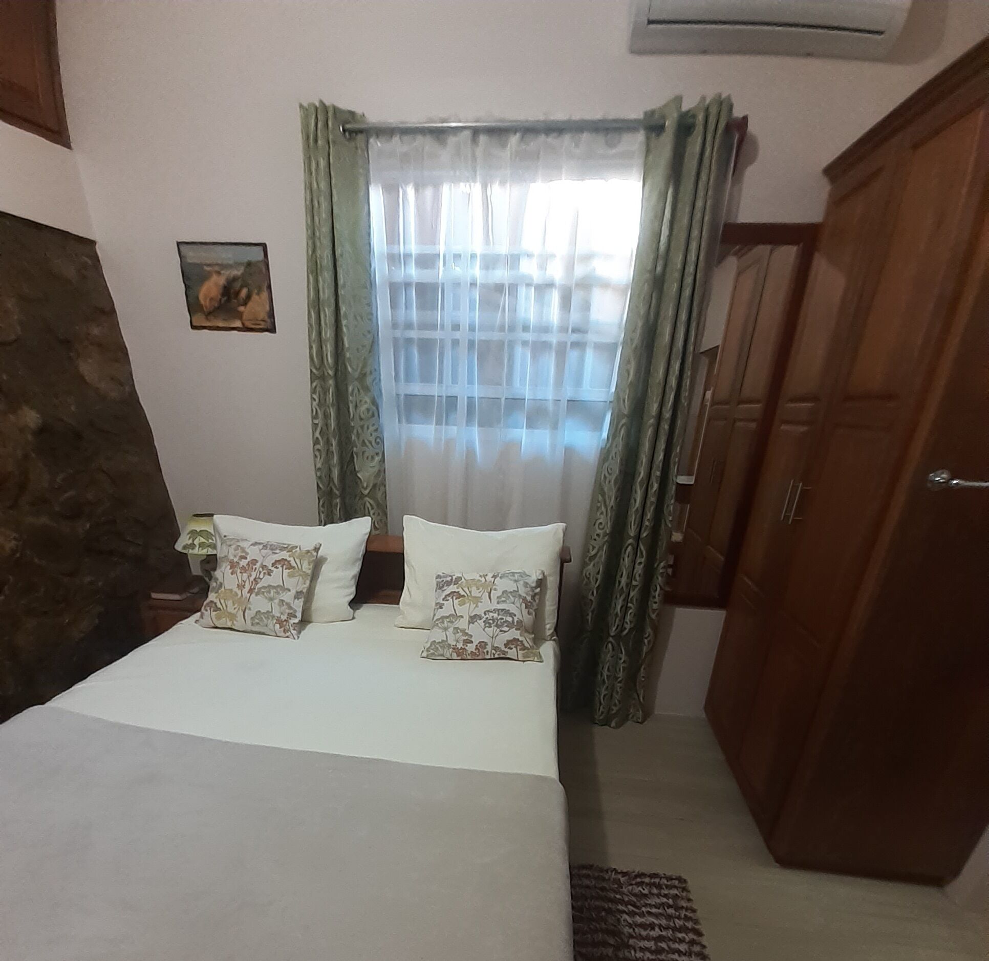 A bedroom with a bed, two pillows, a nightstand, a lamp, a window with curtains, a wall-mounted air conditioner, and a wardrobe.