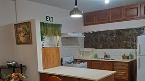 A compact kitchen with wooden cabinets, a white countertop, a gas stove, and a sink. There is a dining table with chairs and a vase of flowers. An 'EXIT' sign is visible on the wall. - Greenhaven Cottage B&B (Les Coteaux)