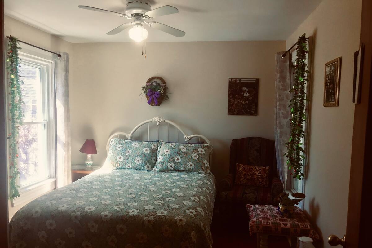 A bedroom with a bed, a ceiling fan, a window with curtains, a bedside table with a lamp, and a chair.