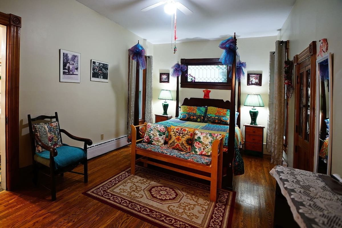 A bedroom with a wooden bed, a chair, a bedside table, a nightstand, a window with curtains, and a ceiling fan.