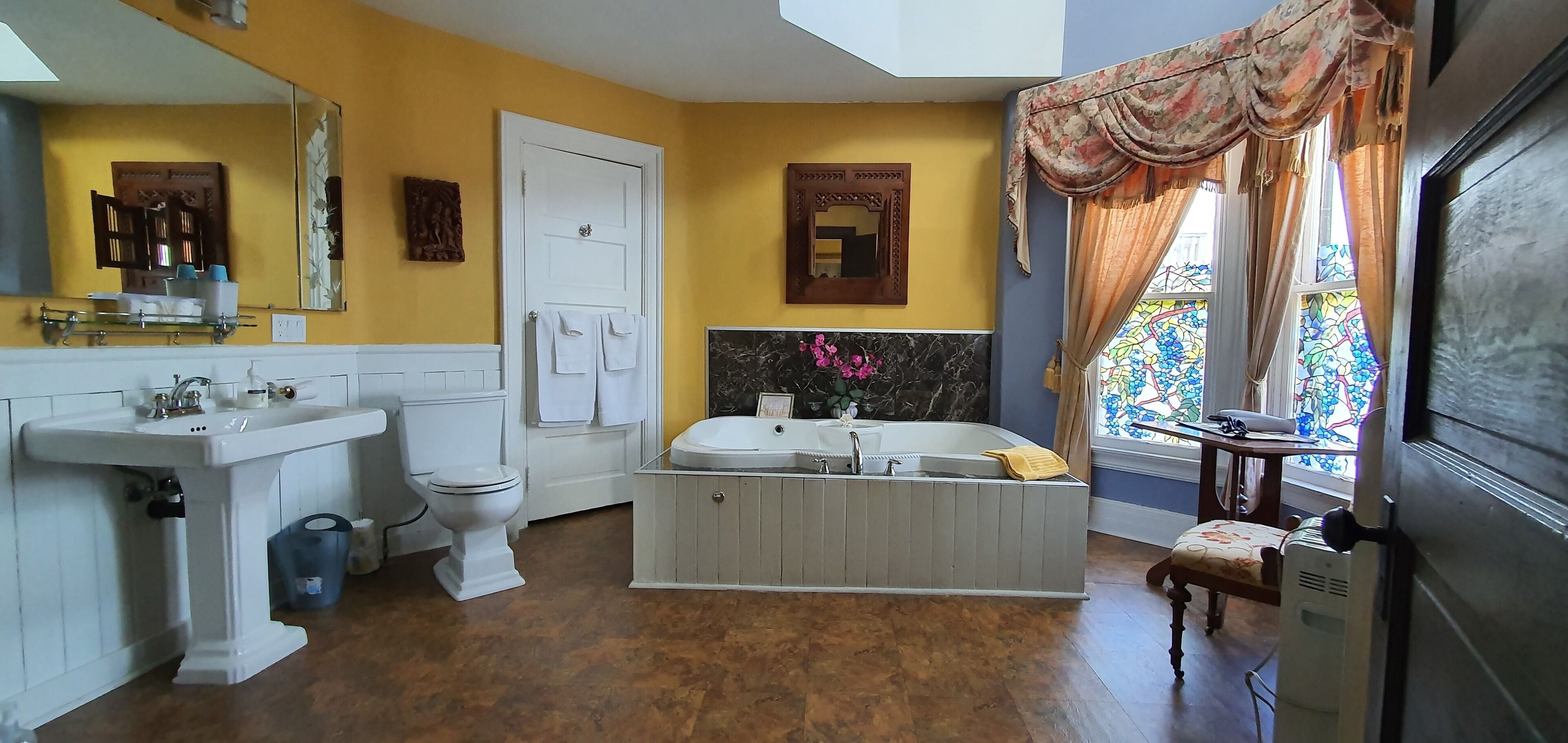 Ein Badezimmer mit Waschbecken, Toilette und Badewanne. Über dem Waschbecken befindet sich ein Spiegel, an der Tür hängt ein Handtuch und es gibt ein Fenster mit Vorhängen.