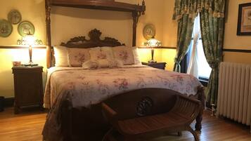Ein traditionell eingerichtetes Schlafzimmer mit Himmelbett, floralem Bettbezug, zwei Nachttischen mit Lampen und einem Holzstuhl.