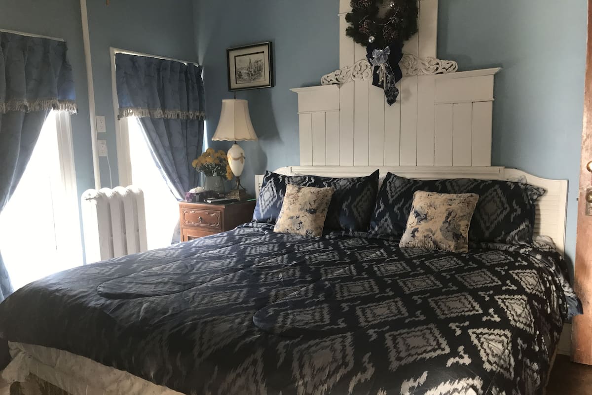 A bedroom with a patterned bedspread, a white headboard, a nightstand with a lamp, and a framed picture on the wall.