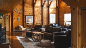A cozy log cabin interior with a wooden ceiling, a fireplace, a comfortable seating area with a sofa and chairs, and a large window providing natural light.