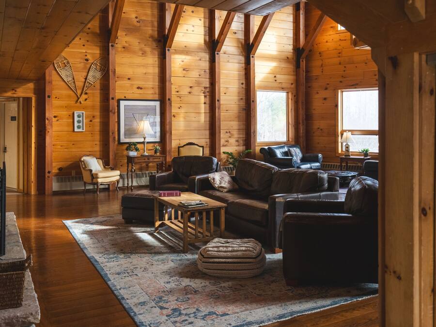 Interior acogedor de una cabaña de madera con techo de madera, chimenea, zona de estar confortable con sofá y sillones, y un ventanal grande que proporciona luz natural.