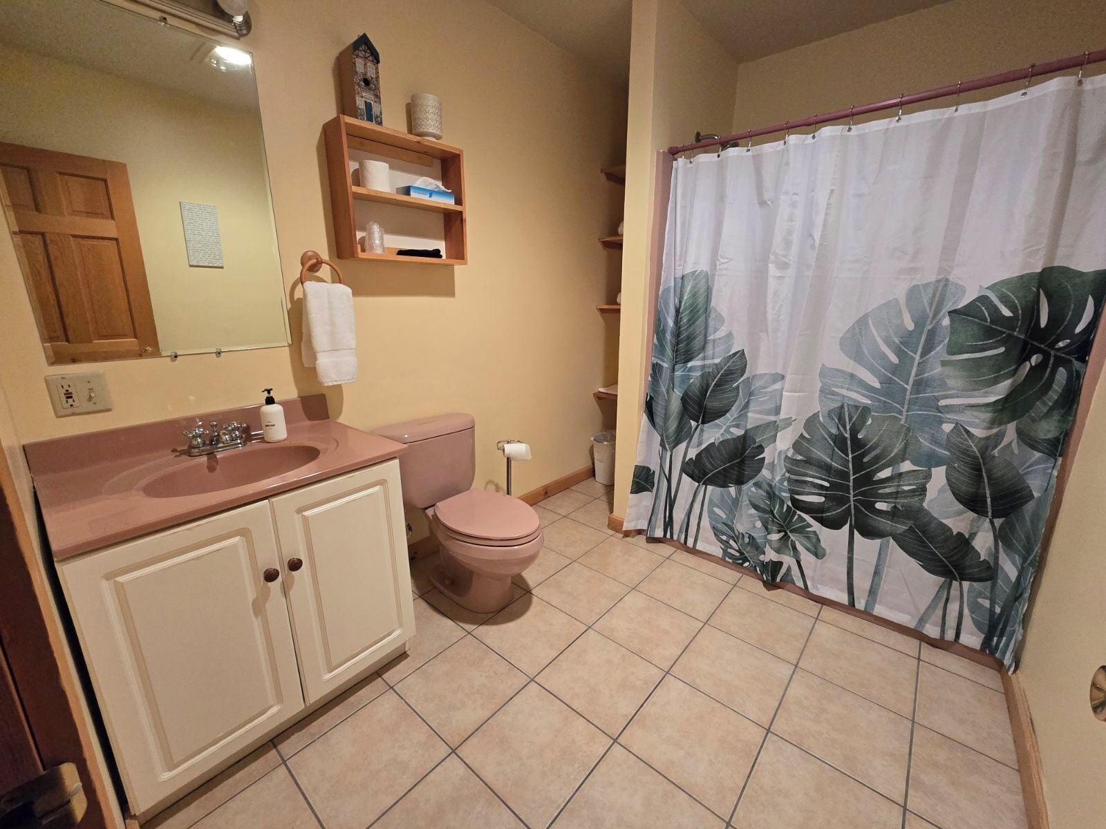 Un baño con lavamanos, inodoro y cortina de ducha rosada con estampado de hojas tropicales.