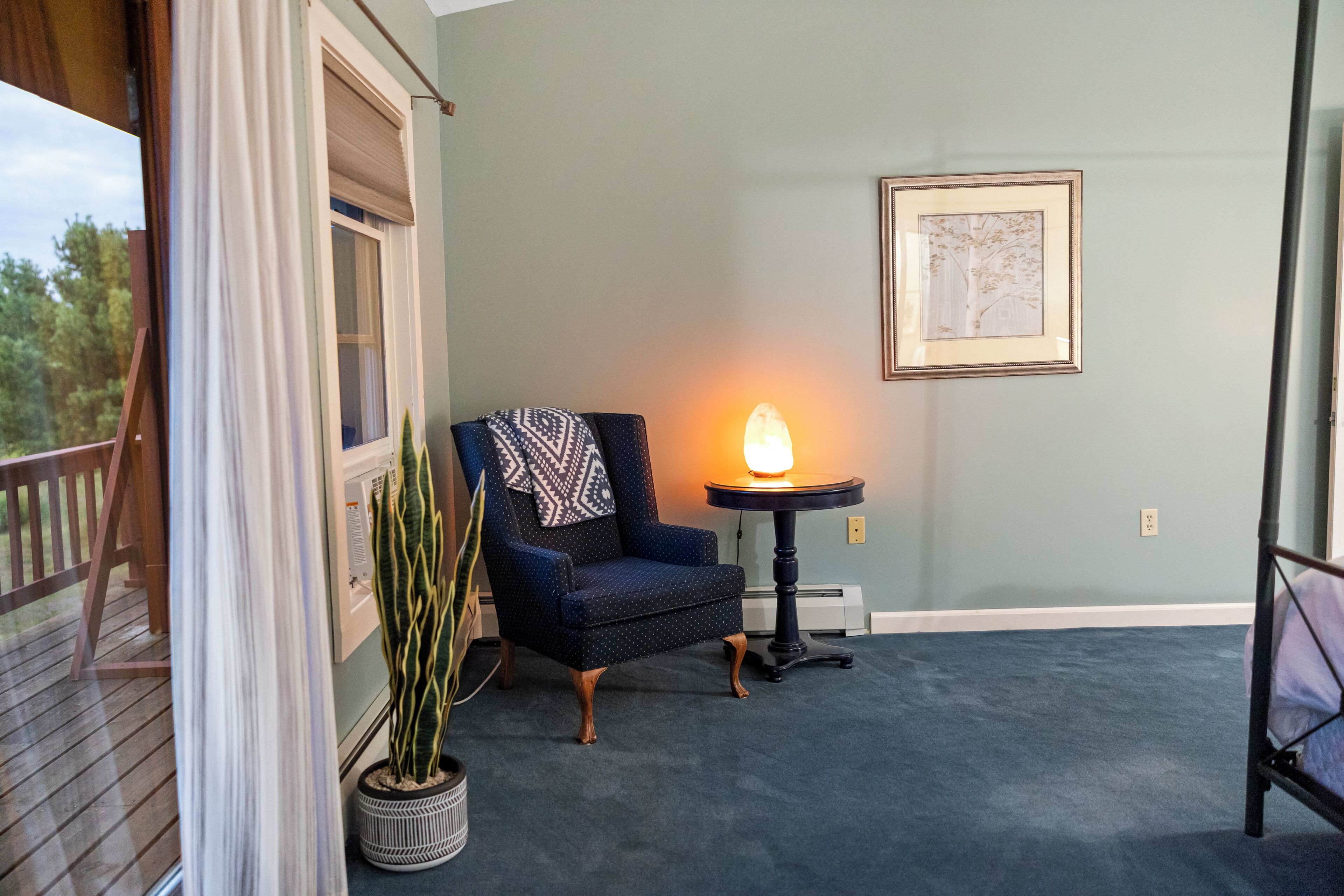 A room with a blue carpet, a chair with a patterned throw, a table with a lamp, a potted plant, and a framed picture on the wall.