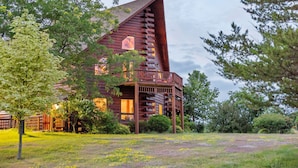 A log cabin with a balcony, surrounded by trees and a grassy field.