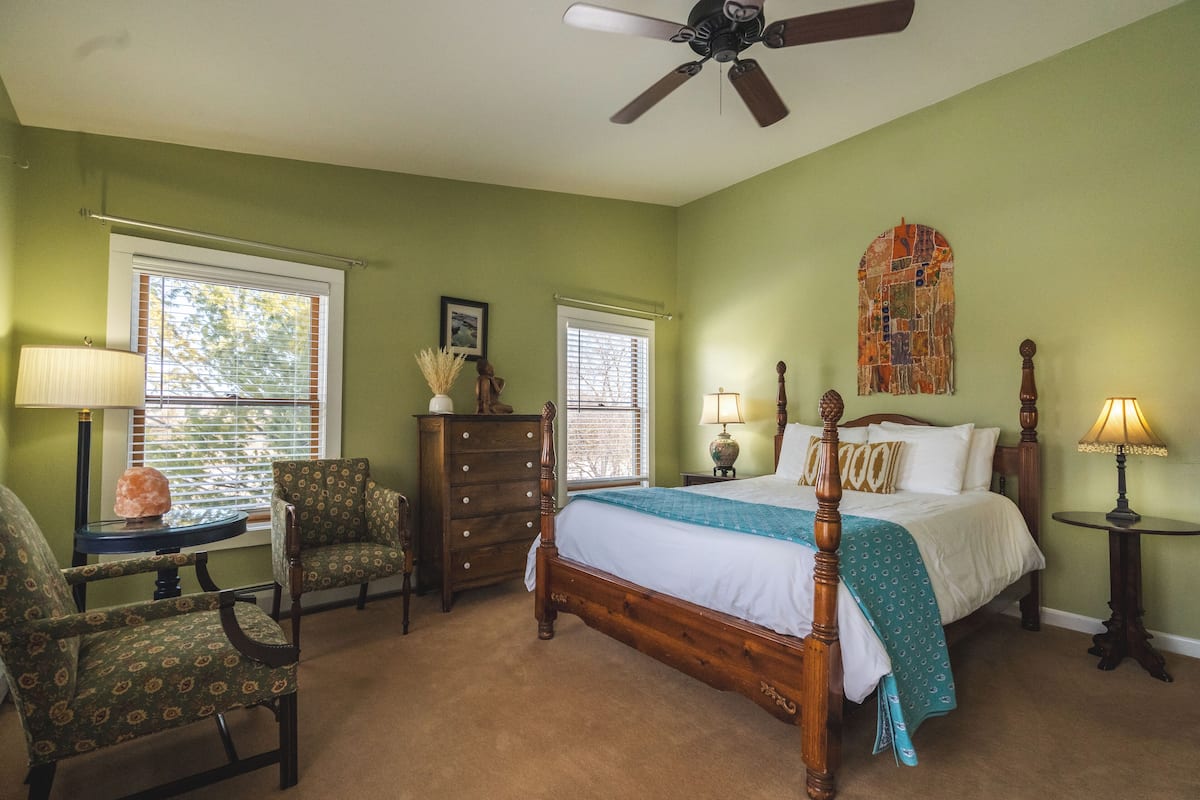 Quarto com cama de madeira, duas cadeiras, cômoda e ventilador de teto.