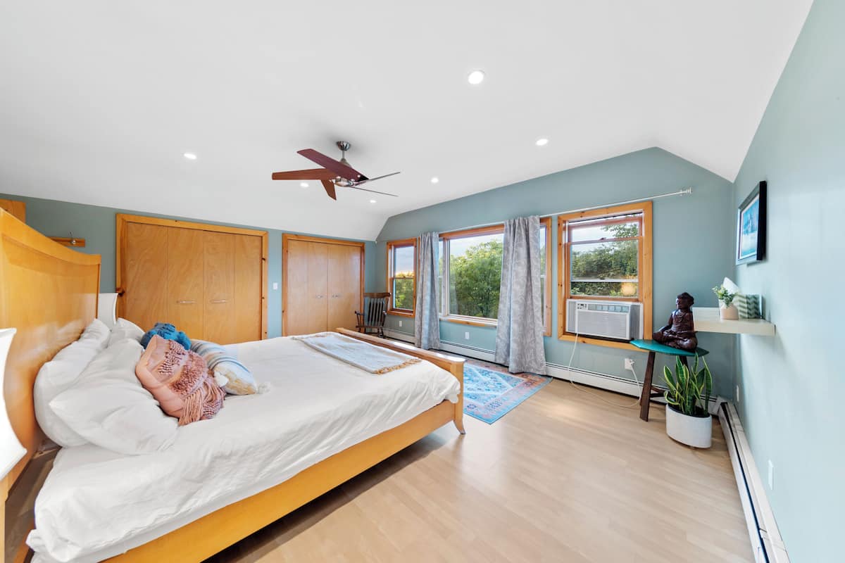 Quarto com cama, guarda-roupa de madeira, ventoinha no teto, janelas amplas e uma pequena mesa com uma planta.