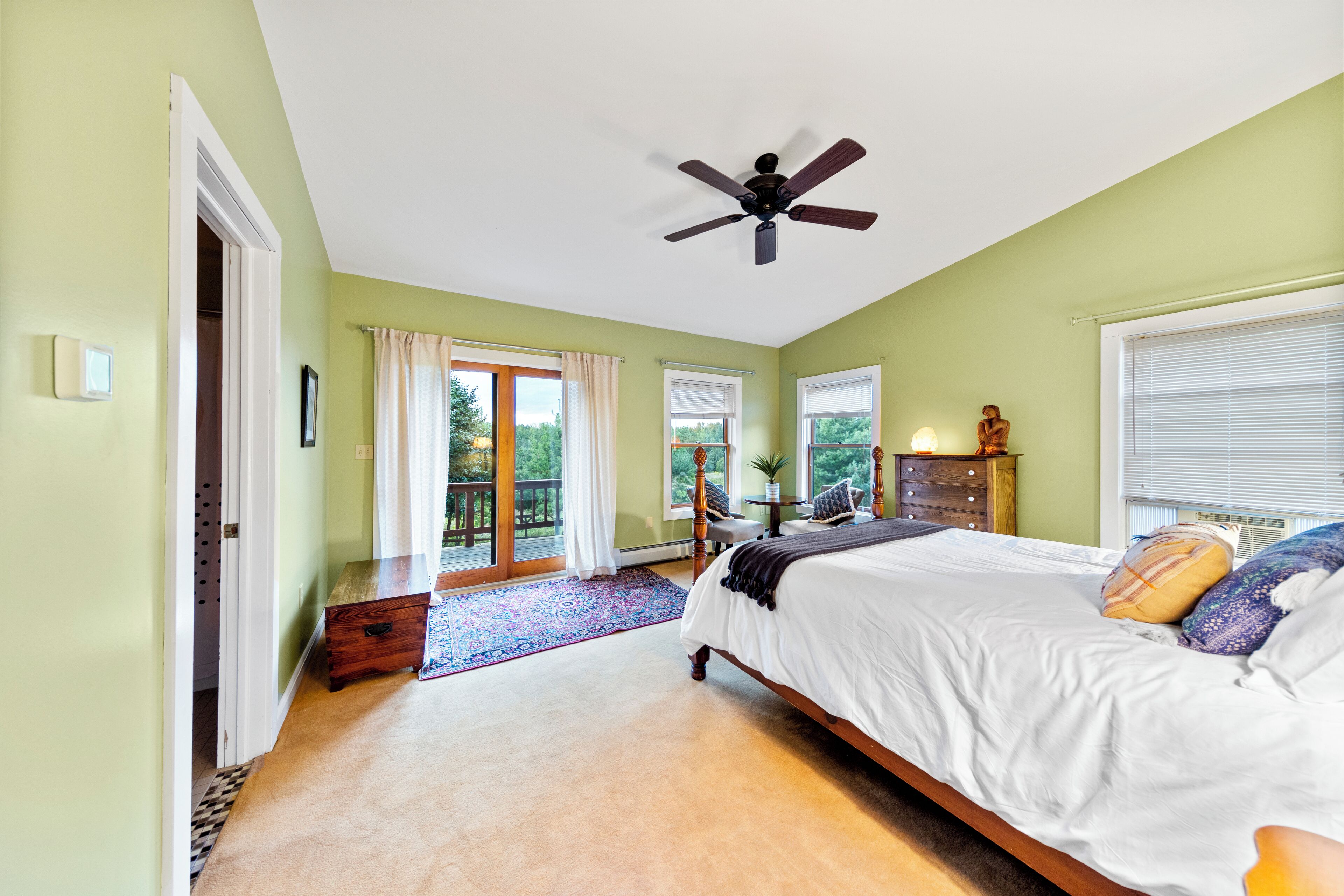 A bedroom with a bed, a ceiling fan, a window with blinds, a door leading to a balcony, and a small table with a plant.