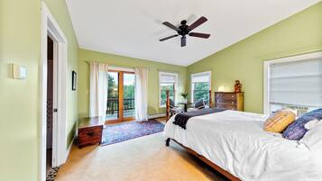 A bedroom with a bed, a ceiling fan, a window with blinds, a door leading to a balcony, and a small table with a plant.