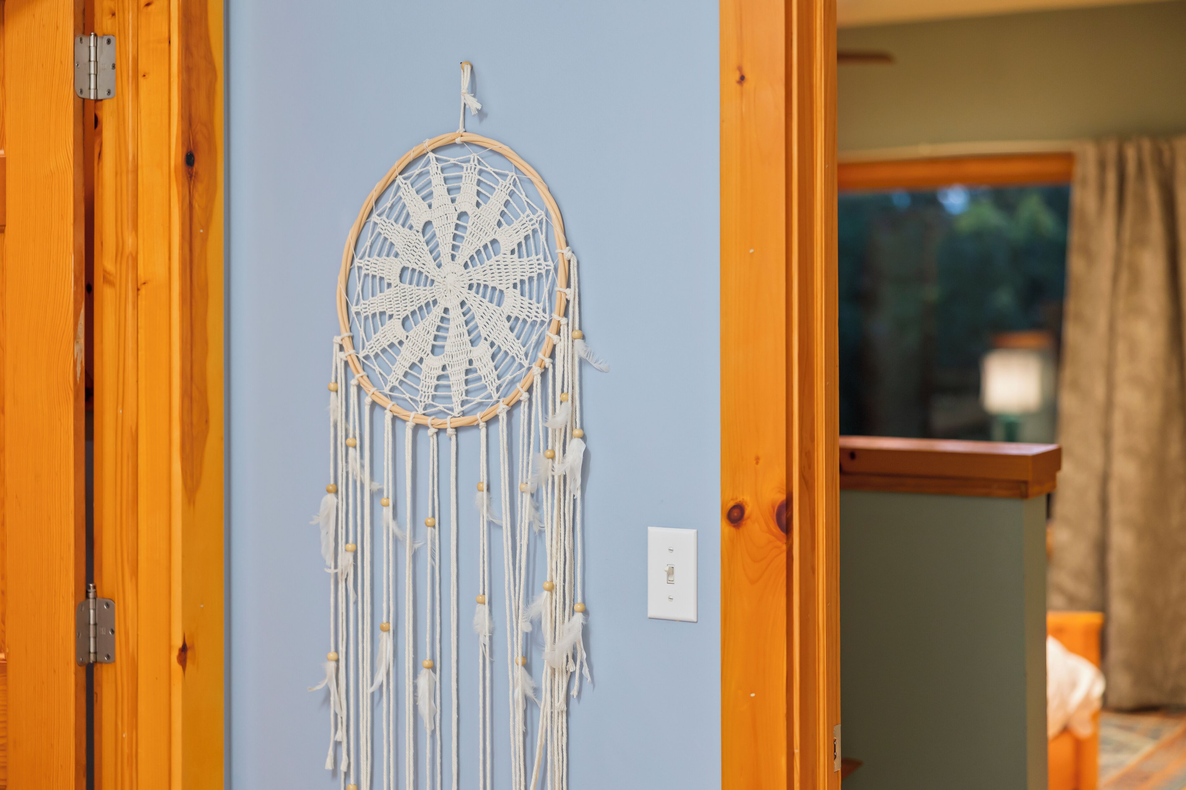 Una habitación con puerta de madera, un atrapasueños colgado en la pared y una ventana con cortinas.