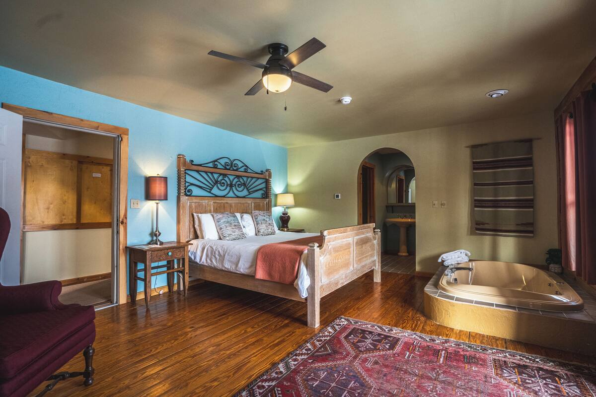 Quarto com cama de madeira, uma poltrona vermelha, uma janela grande, um ventilador de teto e uma banheira.