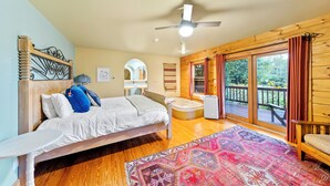 A bedroom with a wooden headboard, a bed, a bedside table, a ceiling fan, a large window with curtains, and a view of the outdoors.