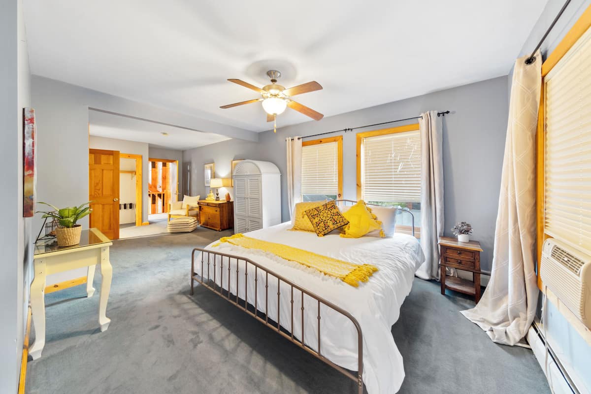 Quarto com cama, ventoinha no teto, mesa de cabeceira, abajur e janela com cortinas.