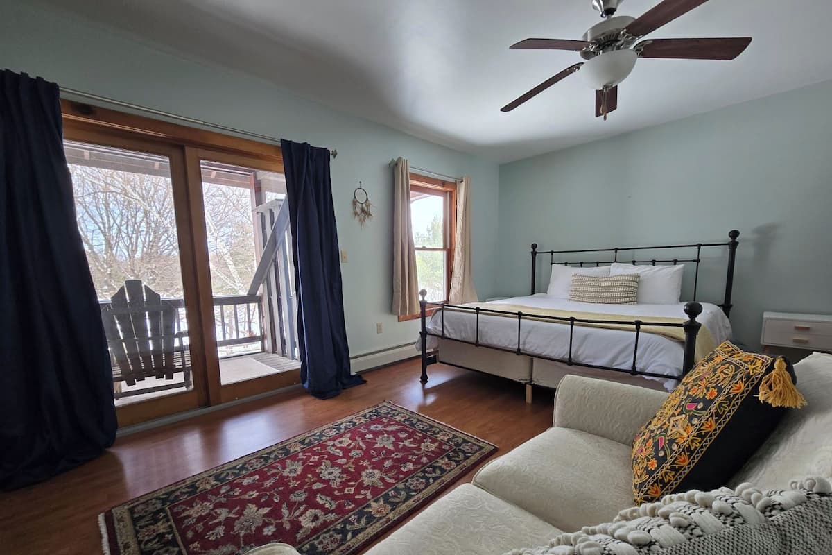 Quarto com cama, ventilador de teto, janela com cortinas, piso de madeira e tapete estampado.