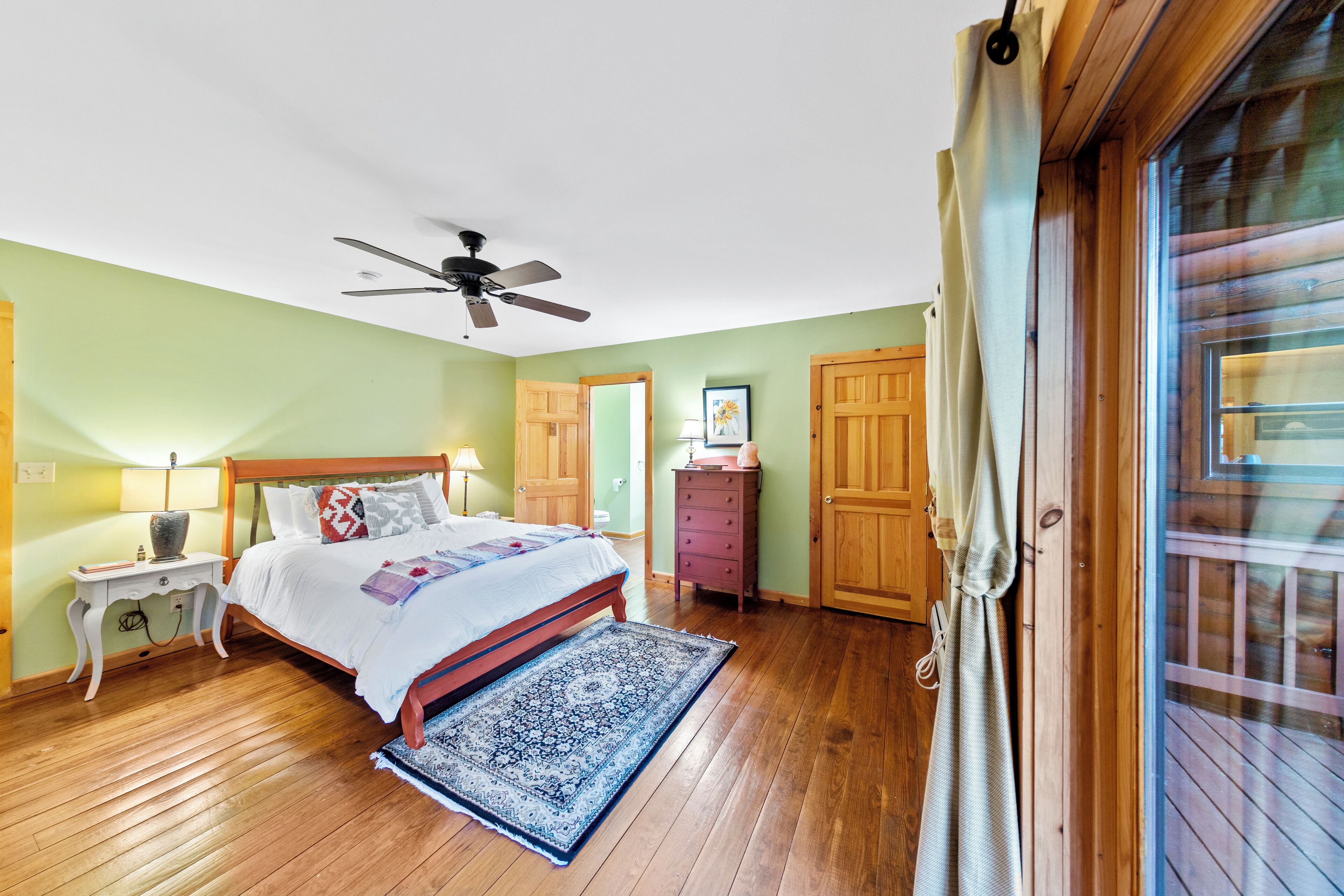 A bedroom with a bed, wooden floor, ceiling fan, and a view of the outside through a door.