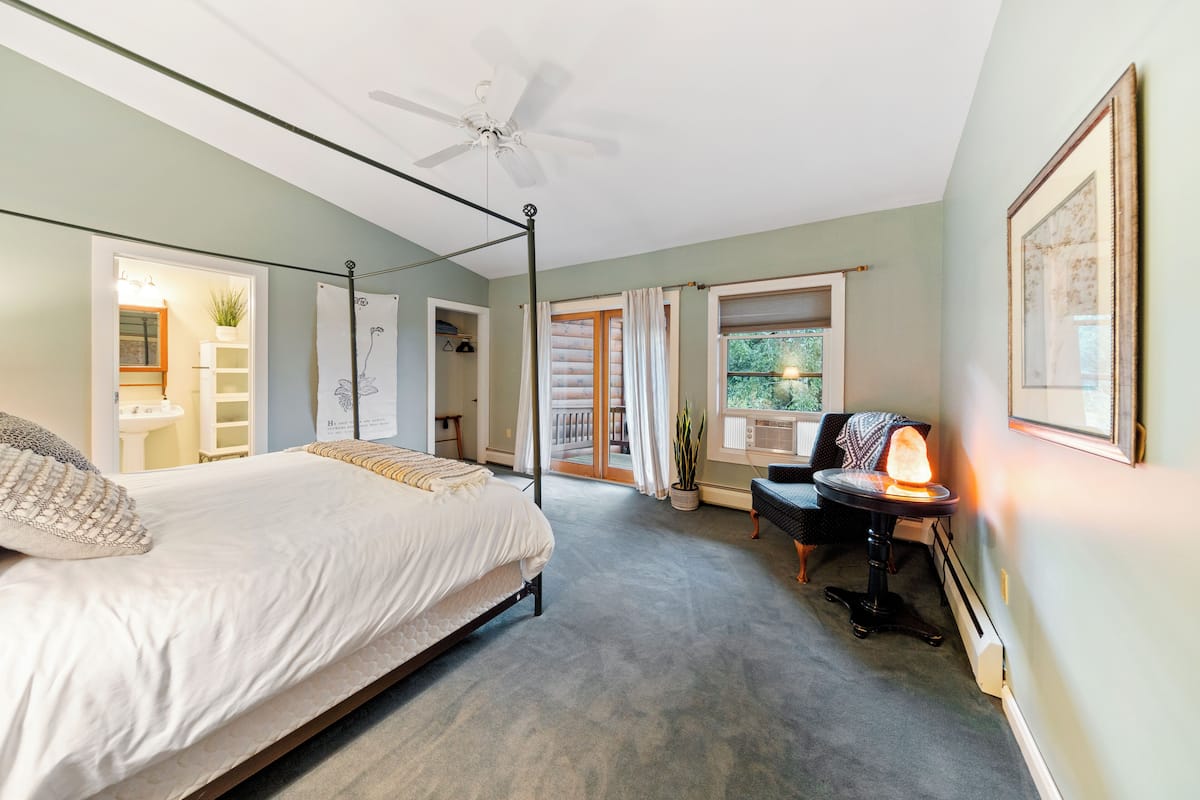 Quarto com cama de dossel, ventoinha no teto, escrivaninha com abajur e vista para o exterior por uma porta deslizante.